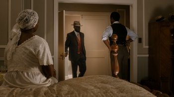 Movie still from “Bessie” (2015), directed by Dee Rees – A man in a suit and a hat standing next to a bed; Medium shot, Over the shoulder angle