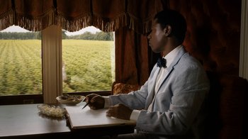 Movie still from “Bessie” (2015), directed by Dee Rees – A man sitting at a table writing on a piece of paper in front of a window; Medium shot, Over the shoulder angle