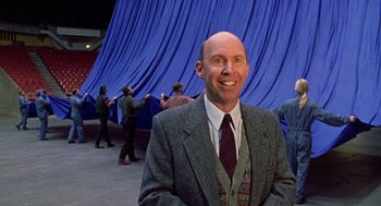Movie still from “Best in Show” (2000), directed by Christopher Guest – A man in a suit and tie standing in front of a blue curtain; Medium shot, Low angle