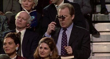 Movie still from “Best in Show” (2000), directed by Christopher Guest – A man in a suit and tie holding a cigarette and wearing sunglasses; Medium shot, Over the shoulder angle