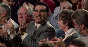 Movie still from “Best in Show” (2000), directed by Christopher Guest – A man in a suit and tie sitting in a crowd of people clapping; Medium shot, Over the shoulder angle
