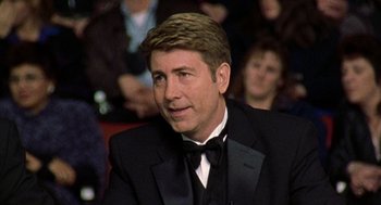 Movie still from “Best in Show” (2000), directed by Christopher Guest – A man in a suit sitting in front of a group of people; Close Up shot, Over the shoulder angle