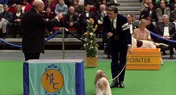Movie still from “Best in Show” (2000), directed by Christopher Guest – A man in a suit and tie standing next to a small dog; Wide shot, Over the shoulder angle