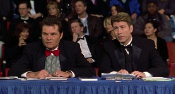 Movie still from “Best in Show” (2000), directed by Christopher Guest – A couple of men sitting at a table in front of a crowd; Medium shot, Low angle