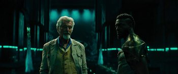 Movie still from “Black Adam” (2022), directed by Jaume Collet-Serra – A man standing next to another man in a dark room; Medium shot, Over the shoulder angle