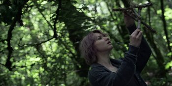 Movie still from “Blair Witch” (2016), directed by Adam Wingard – A woman standing in the middle of a lush green forest; Medium shot, Low angle