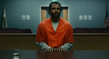 Movie still from “Blindspotting” (2018), directed by Carlos López Estrada – A man in an orange prison uniform sitting at a table in an empty courtroom; Wide shot, Low angle