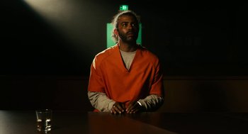 Movie still from “Blindspotting” (2018), directed by Carlos López Estrada – A man in an orange prison uniform sitting at a table in front of an exit sign; Medium shot, Low angle