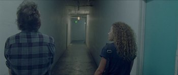 Movie still from “Bliss” (2019), directed by Joe Begos – A woman standing in an empty hallway looking up; Medium shot, Over the shoulder angle