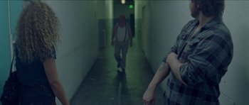Movie still from “Bliss” (2019), directed by Joe Begos – A man walking down a hallway with a person walking by; Medium shot, Over the shoulder angle
