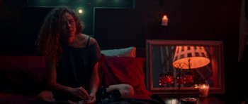 Movie still from “Bliss” (2019), directed by Joe Begos – A woman sitting on a couch next to a fireplace; Medium shot, Over the shoulder angle