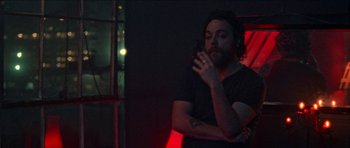 Movie still from “Bliss” (2019), directed by Joe Begos – A bearded man smoking a cigarette in a dark room; Medium shot, Over the shoulder angle