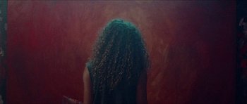 Movie still from “Bliss” (2019), directed by Joe Begos – A woman with long curly hair sitting in front of a red wall; Medium shot, Overhead angle
