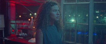 Movie still from “Bliss” (2019), directed by Joe Begos – A woman with curly hair standing next to a window at night; Medium shot, Over the shoulder angle