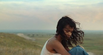 Movie still from “Bones and All” (2022), directed by Luca Guadagnino – A woman sitting on top of a grass covered field; Medium shot, Over the shoulder angle