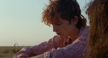 Movie still from “Bones and All” (2022), directed by Luca Guadagnino – A young man with long hair is sitting on a beach; Close Up shot, Over the shoulder angle