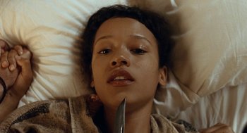 Movie still from “Bones and All” (2022), directed by Luca Guadagnino – A woman laying in a bed with a knife in her mouth; Close Up shot, Overhead angle