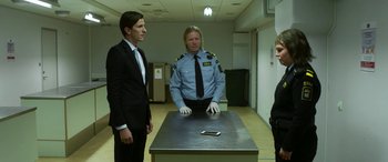 Movie still from “Border” (2018), directed by Ali Abbasi – Two men in suits stand next to a table; Medium shot, Low angle