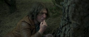 Movie still from “Border” (2018), directed by Ali Abbasi – A man sitting in the grass eating something off of a tree; Close Up shot, High angle