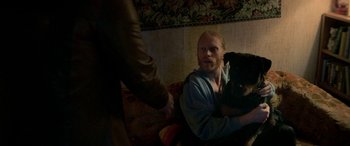 Movie still from “Border” (2018), directed by Ali Abbasi – A man sitting on a couch holding a black dog; Medium shot, Over the shoulder angle