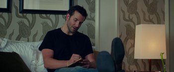 Movie still from “Burnt” (2015), directed by John Wells – A man sitting on a couch writing on a notebook; Medium shot, Over the shoulder angle