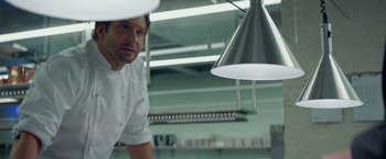 Movie still from “Burnt” (2015), directed by John Wells – A man standing in front of some lights in a kitchen; Medium shot, Low angle