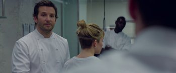 Movie still from “Burnt” (2015), directed by John Wells – A group of people standing around a woman in a room; Close Up shot, Over the shoulder angle