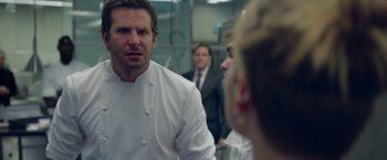 Movie still from “Burnt” (2015), directed by John Wells – A group of people in a kitchen talking to each other; Close Up shot, Over the shoulder angle