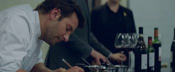 Movie still from “Burnt” (2015), directed by John Wells – A man writing on a sheet of paper next to a wine glass; Close Up shot, Over the shoulder angle
