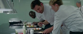 Movie still from “Burnt” (2015), directed by John Wells – A group of people standing around a table with plates of food on top of it; Medium shot, Over the shoulder angle
