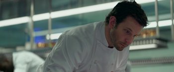 Movie still from “Burnt” (2015), directed by John Wells – A man wearing a white chef's coat and a black hat; Close Up shot, Over the shoulder angle