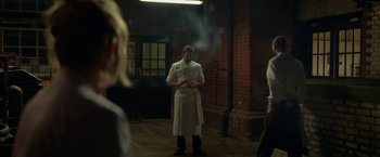 Movie still from “Burnt” (2015), directed by John Wells – A man standing in an industrial setting with smoke coming out of his mouth; Medium shot, Over the shoulder angle