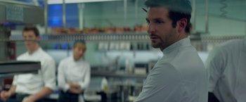 Movie still from “Burnt” (2015), directed by John Wells – A man in a white dress shirt is looking at the camera while another man in a white dress shirt is sitting in the background; Close Up shot, Over the shoulder angle