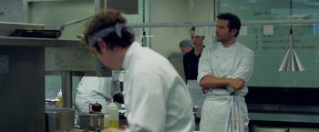 Movie still from “Burnt” (2015), directed by John Wells – A group of people standing in a kitchen preparing food; Medium shot, Over the shoulder angle