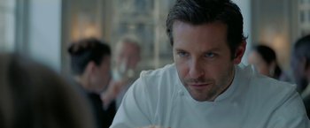 Movie still from “Burnt” (2015), directed by John Wells – A man in a chef's outfit sitting at a table; Close Up shot, Over the shoulder angle