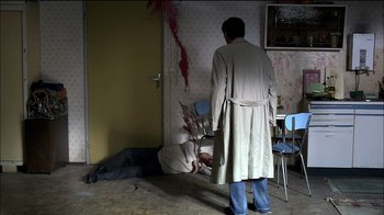 Movie still from “Caché” (2005), directed by Michael Haneke – A man in a trench coat standing in front of a man lying on the floor; Wide shot, High angle