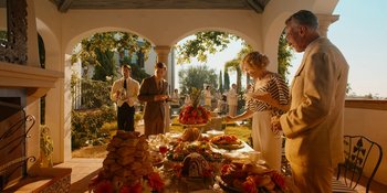 Movie still from “Café Society” (2016), directed by Woody Allen – A group of people standing in front of a table filled with food; Wide shot, Over the shoulder angle