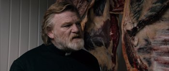 Movie still from “Calvary” (2014), directed by John Michael McDonagh – A man with a long beard and a beard stand in front of a wall of meat; Medium shot, Low angle