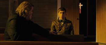 Movie still from “Calvary” (2014), directed by John Michael McDonagh – A man sitting in front of another man in front of a wooden wall; Medium shot, Over the shoulder angle