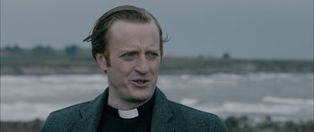 Movie still from “Calvary” (2014), directed by John Michael McDonagh – A man in a priest's outfit standing in front of a body of water; Close Up shot, Over the shoulder angle