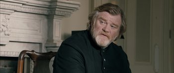Movie still from “Calvary” (2014), directed by John Michael McDonagh – A man with a beard and a black shirt is sitting in front of a fireplace; Close Up shot, Over the shoulder angle