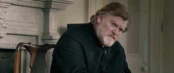 Movie still from “Calvary” (2014), directed by John Michael McDonagh – A man with long hair and a goatee sitting in front of a chair; Close Up shot, Low angle