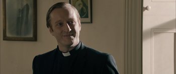 Movie still from “Calvary” (2014), directed by John Michael McDonagh – A man wearing a priest's outfit and smiling for the camera; Close Up shot, Over the shoulder angle