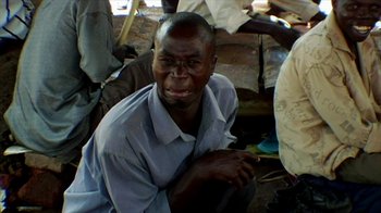 Movie still from “Cameraperson” (2016), directed by Kirsten Johnson – A man sitting on the ground in front of other people; Close Up shot, High angle