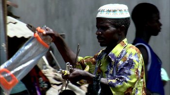 Movie still from “Cameraperson” (2016), directed by Kirsten Johnson – A man in a blue and yellow shirt holding a pair of scissors; Medium shot, Low angle