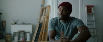 Movie still from “Candyman” (2021), directed by Nia DaCosta – A man wearing a red hat sitting in front of a painting; Close Up shot, Over the shoulder angle
