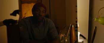 Movie still from “Candyman” (2021), directed by Nia DaCosta – A man sitting in front of a table with a bottle; Close Up shot, Over the shoulder angle