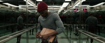 Movie still from “Candyman” (2021), directed by Nia DaCosta – A man in a gray shirt and a red hat is holding a brown bag; Medium shot, Overhead angle