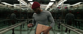 Movie still from “Candyman” (2021), directed by Nia DaCosta – A man in a gray shirt and a red hat is holding papers; Medium shot, Low angle