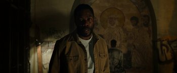 Movie still from “Candyman” (2021), directed by Nia DaCosta – A man standing in front of a wall with a painting behind him; Medium shot, Low angle